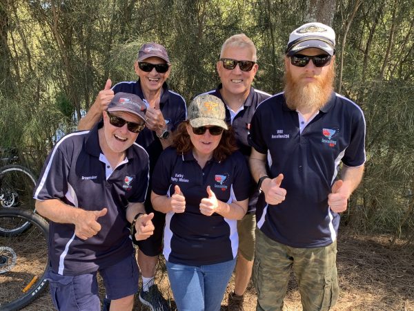 Group of five people standing in a group wearing Geocaching NSW polo shirts, sunglasses and hats, all smiling with their thumbs up.